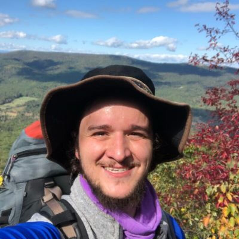 The image shows a man with a beard and a hat, wearing a backpack, likely hiking in a mountainous area. He is smiling at the camera. The background features a scenic view of mountains and trees, with a partly cloudy sky. The overall impression is that he is enjoying an outdoor adventure.